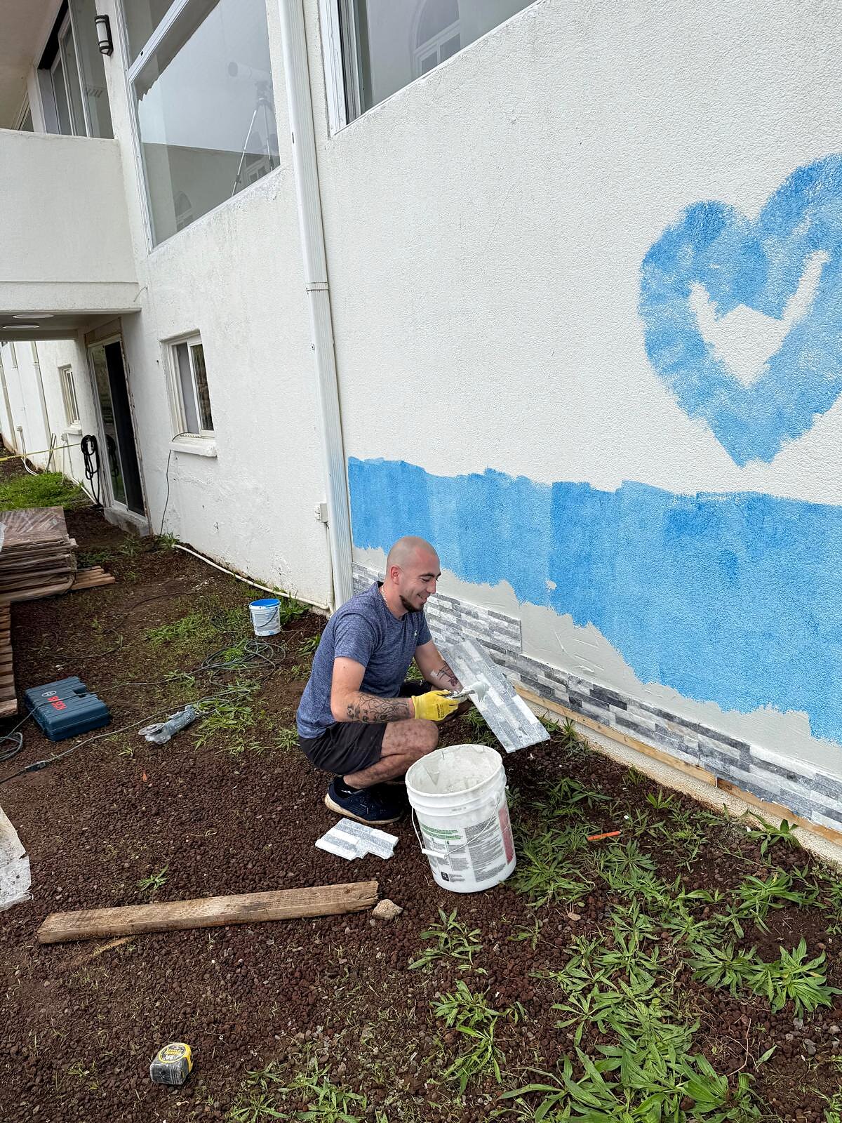 Painter finishing a stone veneer accent wall below a stucco exterior painted blue with a heart design