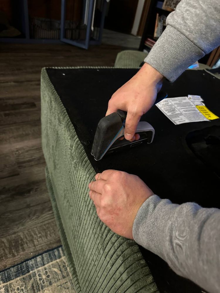 Securing the fabric base of a green sofa with a staple gun during furniture assembly