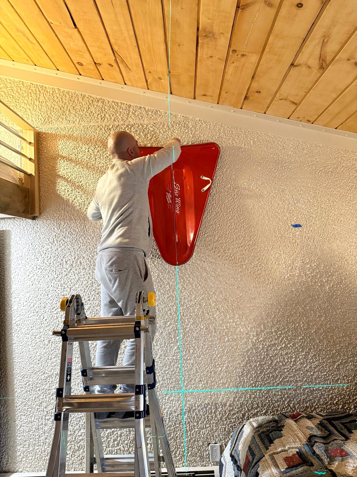 Installer on a ladder mounts a red vintage sled as wall decor on textured stucco, using a laser level to guide placement