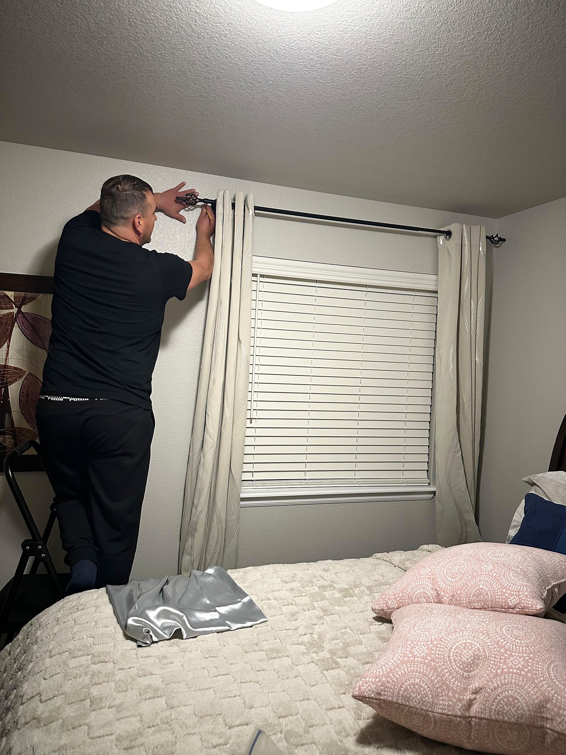 Installer mounting a black curtain rod bracket above a bedroom window with cream curtains