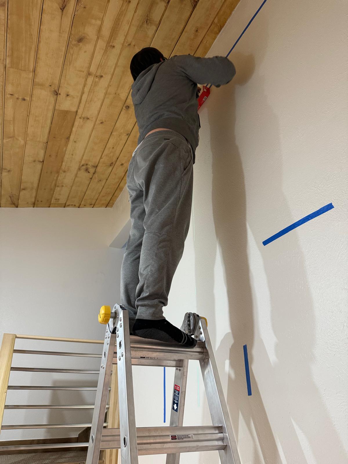 Technician on a ladder marking anchor points on a high stairwell wall below a wood plank ceiling