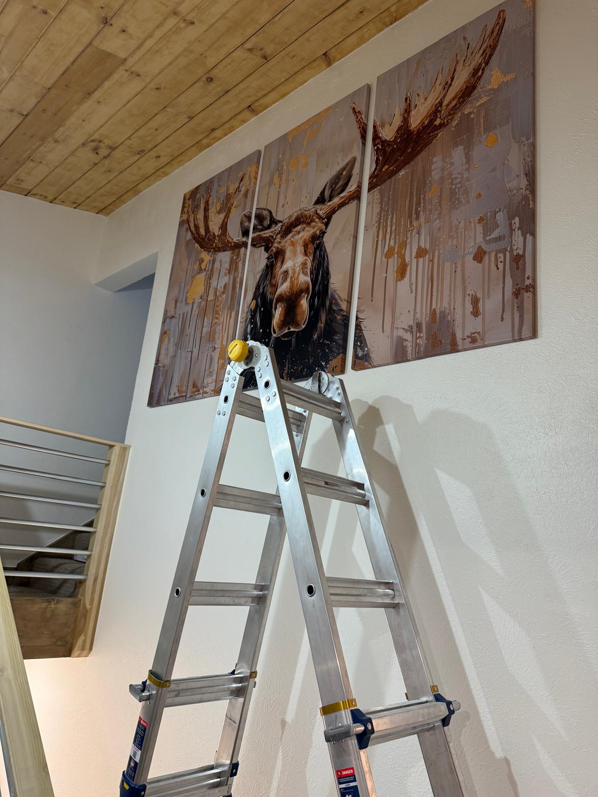 Three-panel moose artwork hung level on a high stairwell wall with a ladder in the foreground