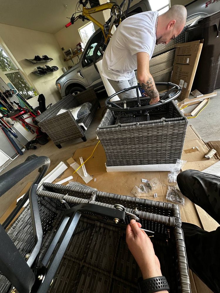 Two technicians assembling wicker patio furniture frames on cardboard in a garage