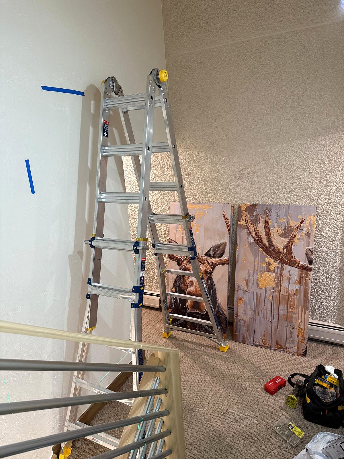 Two-panel gold and brown moose canvas art staged against the wall, ladder set up near the stairway before hanging