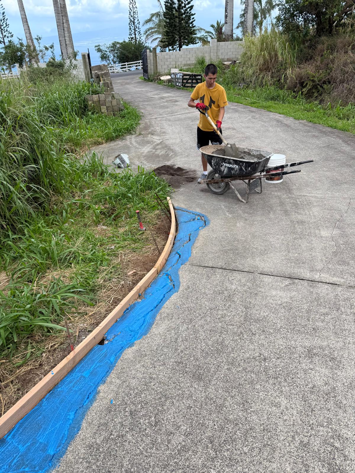 Technician mixing concrete in a wheelbarrow beside a formed driveway curb with the edge sealed in blue membrane