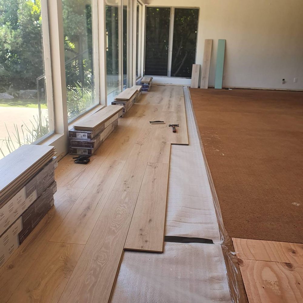Wide-plank laminate flooring being laid over underlayment beside a wall of windows