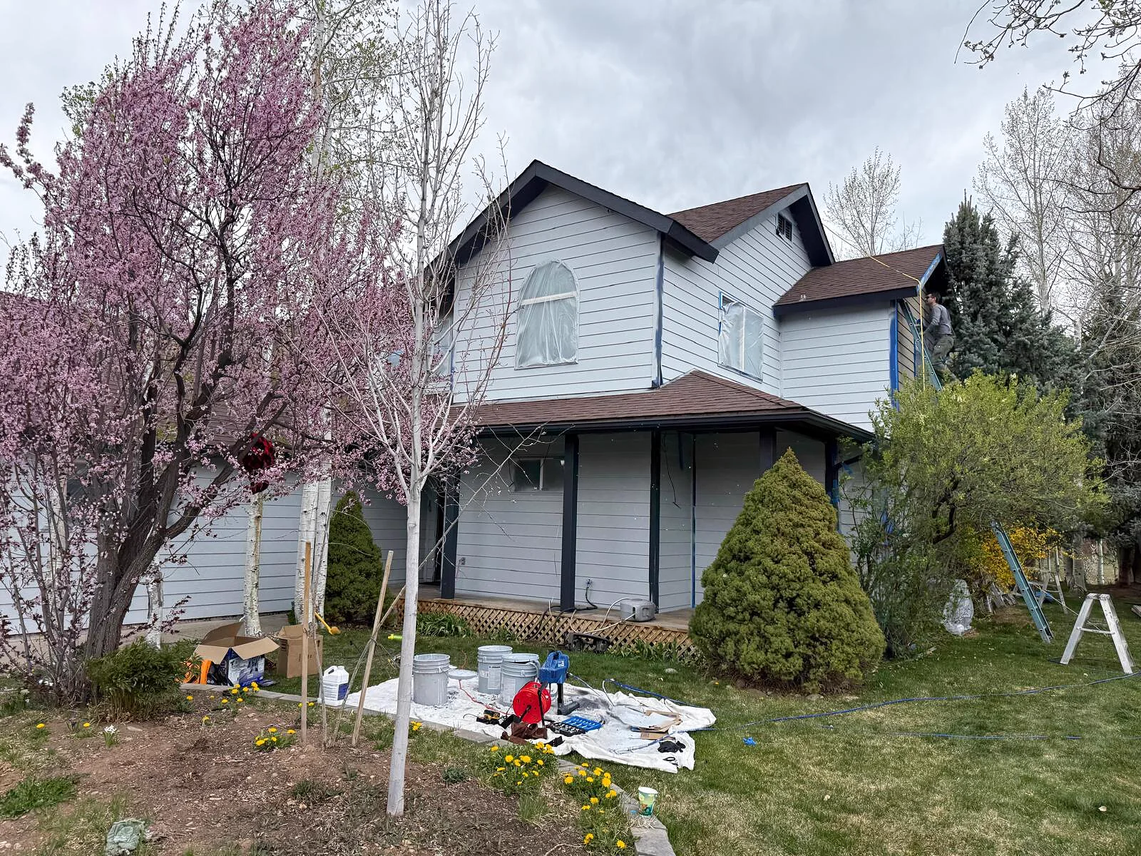 Two-story mountain-area home getting a full exterior repaint, painter working from a ladder near the roofline
