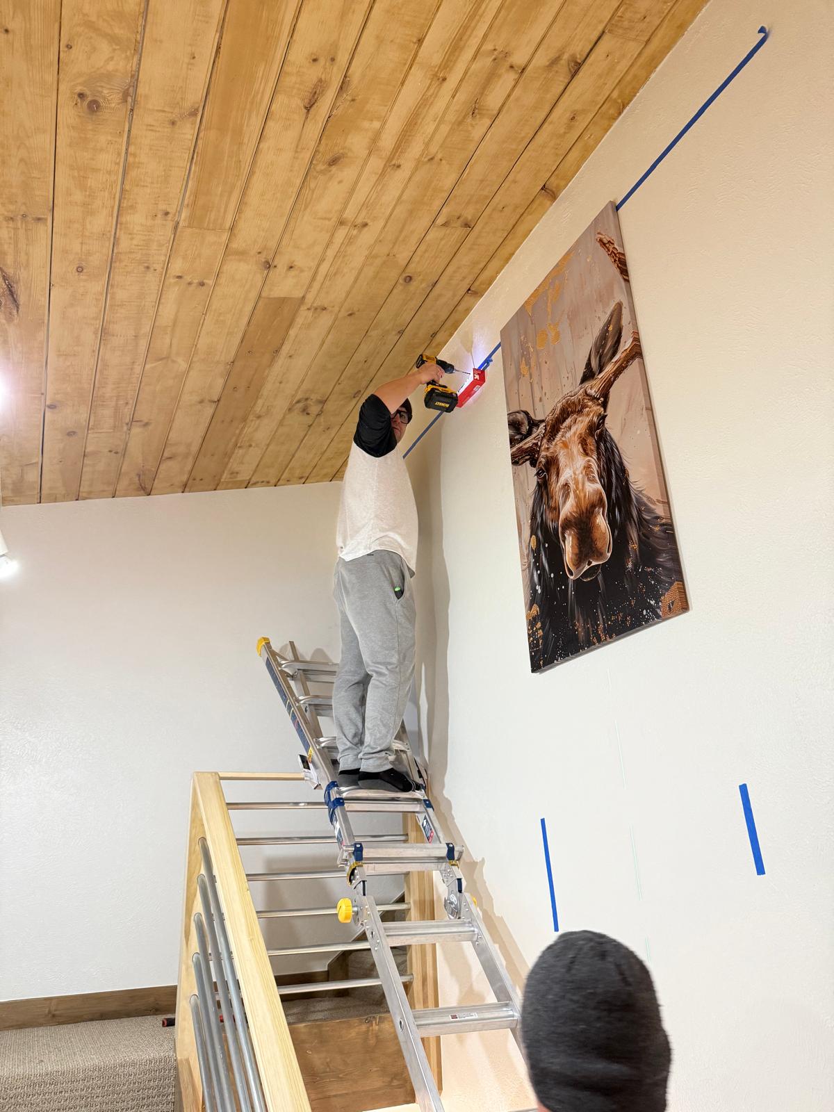 Technician on an extension ladder drilling anchors beside a hung moose canvas panel on a tall wall